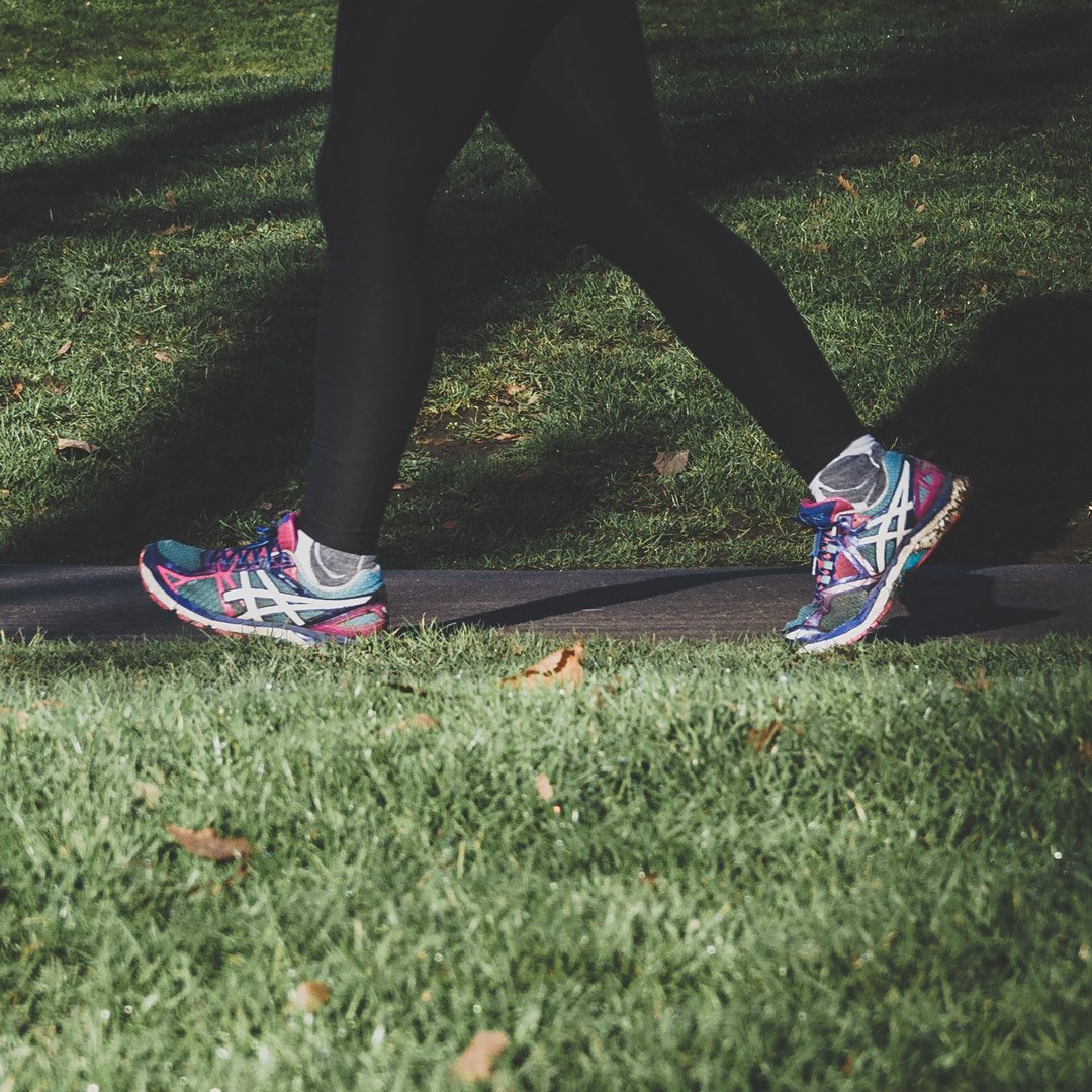 An image of someone walking through a park on a sunny day. Only the bottom half of their legs are visible wearing trainers and leggings